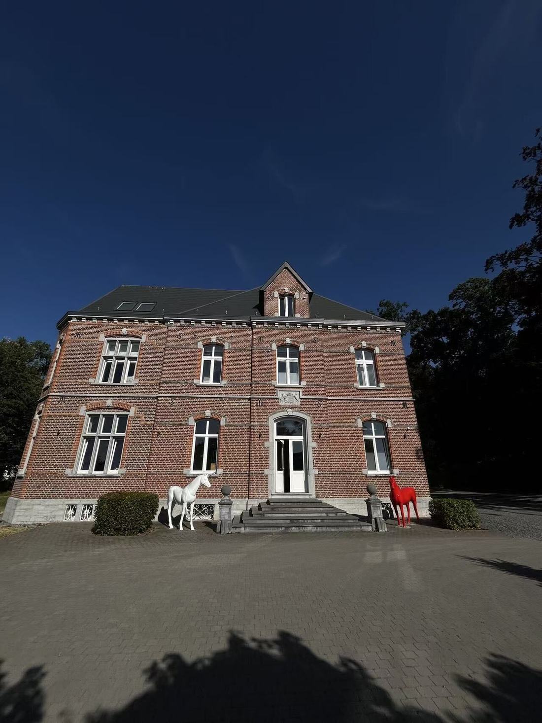 The Clinique de la Brisée building in Maisières, Belgium: a two-storey nineteenth-century red-brick manor with a slate roof, reached by a stone staircase flanked by a white and a red horse sculpture.