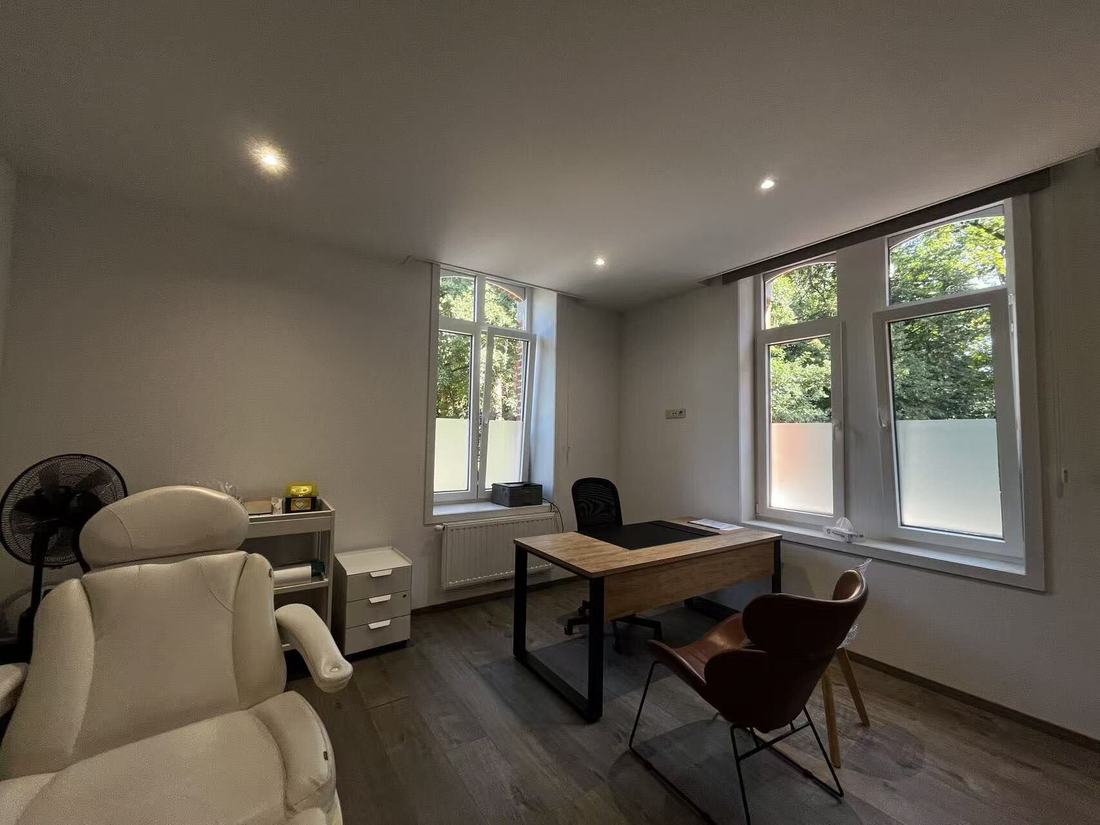 A consulting room with two tall windows onto trees, a wooden desk, an office chair, a visitor's chair and a white reclining examination chair.